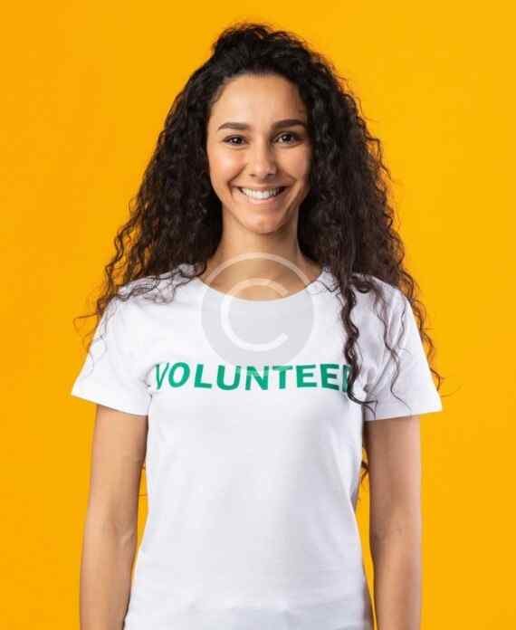 صورة مخزون بعلامة مائية لامرأة مبتسمة بشعر مجعد ترتدي قميصًا أبيض كُتب عليه Volunteer على خلفية صفراء | A watermarked stock image of a smiling woman with curly hair wearing a white T-shirt reading Volunteer against a yellow background