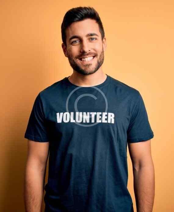 صورة مخزون بعلامة مائية لرجل مبتسم بلحية خفيفة يرتدي قميصًا كحلي اللون كُتب عليه Volunteer على خلفية برتقالية | A watermarked stock image of a smiling man with light stubble wearing a navy T-shirt reading Volunteer against an orange background