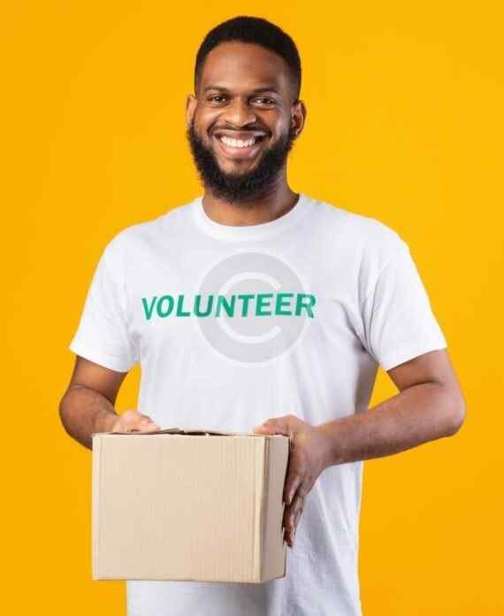 صورة مخزون بعلامة مائية لرجل مبتسم يرتدي قميصًا كُتب عليه Volunteer ويحمل صندوق كرتون على خلفية صفراء | A watermarked stock image of a smiling man wearing a shirt reading Volunteer and holding a cardboard box against a yellow background