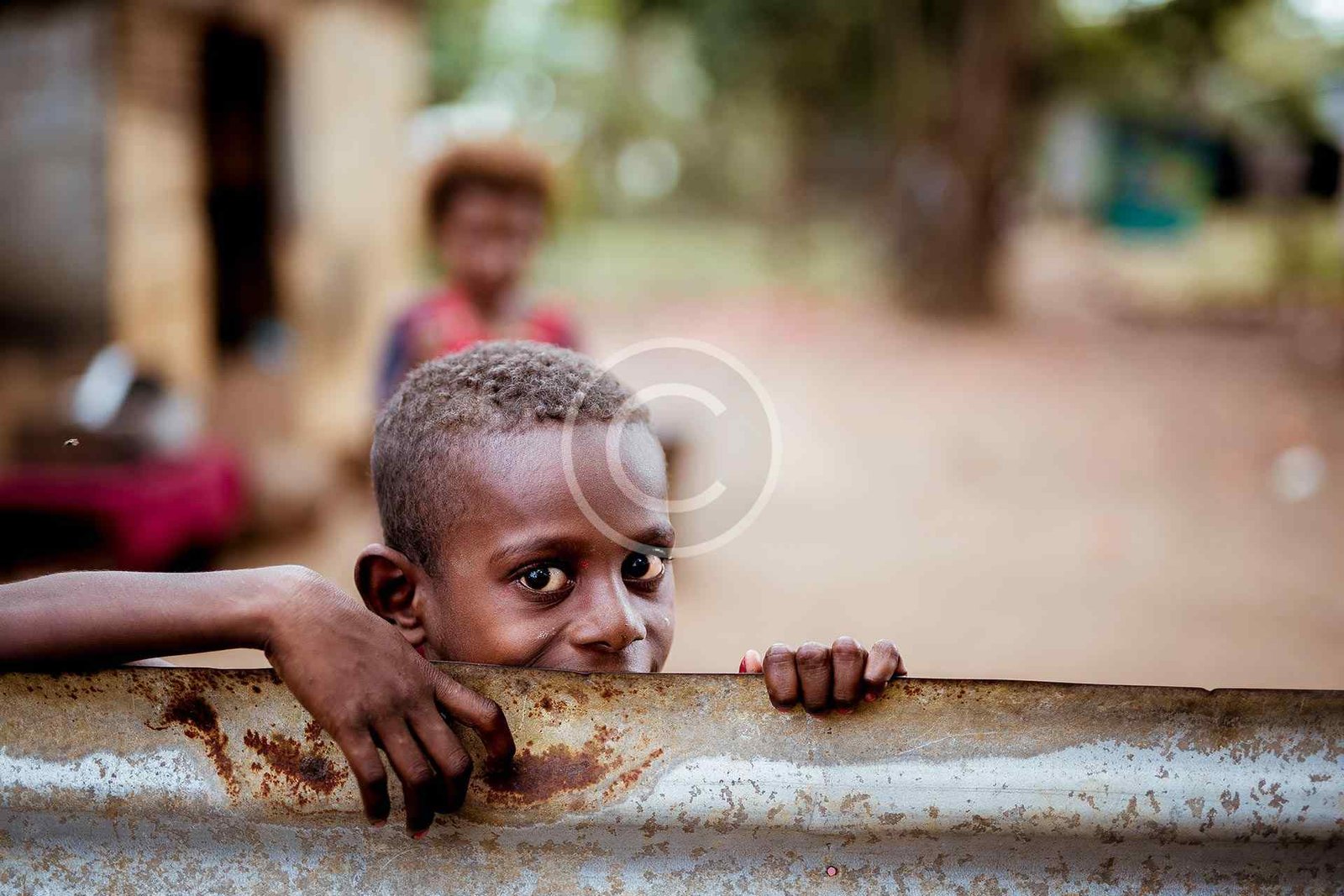 صورة مخزون محمية بحقوق النشر لصبي يطل برأسه فوق سياج معدني صدئ في قرية | Copyright-watermarked stock photo of a boy peeking his head over a rusty metal fence in a village
