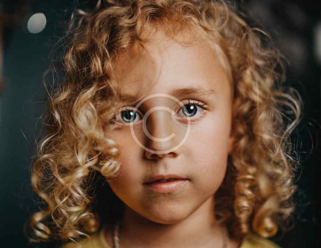 صورة مخزنة محمية بحقوق نشر لوجه طفلة ذات شعر مجعد وعينين فاتحتي اللون بعدسة مقربة | Copyright-watermarked stock close-up of a curly-haired child's face with light-colored eyes