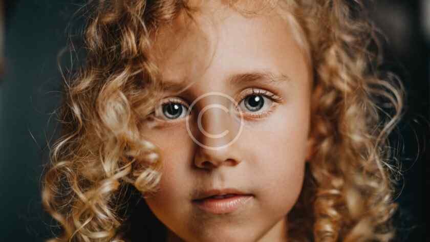صورة مخزنة محمية بحقوق نشر لوجه طفلة ذات شعر مجعد وعينين فاتحتي اللون بعدسة مقربة | Copyright-watermarked stock close-up of a curly-haired child's face with light-colored eyes