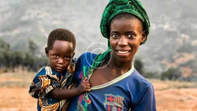 صورة مخزنة محمية بحقوق نشر لمربّية تحمل طفلاً أمام تلال ريفية | Copyright-watermarked stock photo of a woman carrying a child in front of rural hills