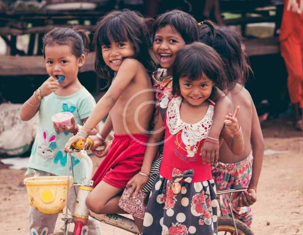 صورة مخزنة محمية بحقوق نشر لمجموعة أطفال مبتسمين يتشاركون دراجة في قرية | Copyright-watermarked stock photo of a group of smiling children sharing a bicycle in a village setting