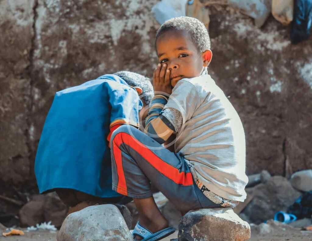 صورة مخزنة محمية بحقوق نشر لصبي جالس على صخور مع طفل آخر أمام جدار طيني | Copyright-watermarked stock photo of a boy sitting on rocks beside another child in front of a mud wall