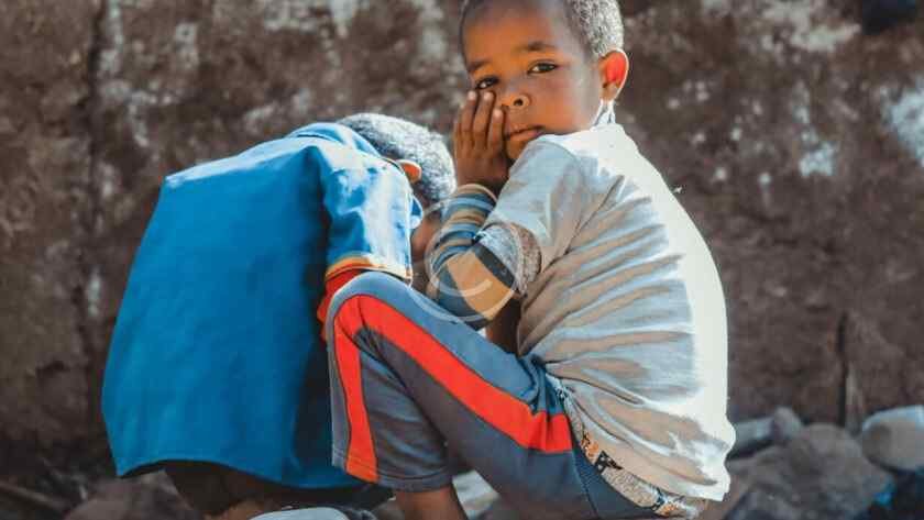 صورة مخزنة محمية بحقوق نشر لصبي جالس على صخور مع طفل آخر أمام جدار طيني | Copyright-watermarked stock photo of a boy sitting on rocks beside another child in front of a mud wall