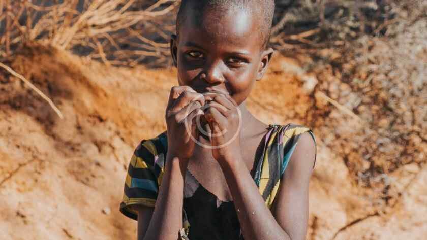 صورة مخزنة محمية بحقوق نشر لطفل يغطي فمه بيديه أمام خلفية أرض جافة قاحلة | Copyright-watermarked stock photo of a child covering his mouth with both hands against an arid, barren backdrop