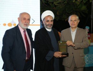 ثلاثة رجال بينهم رجل دين بعمامة بيضاء يمسكون درعاً تكريمياً أمام شاشة عرض ملونة | Three men including a cleric in a white turban hold a commemorative plaque before a colourful screen