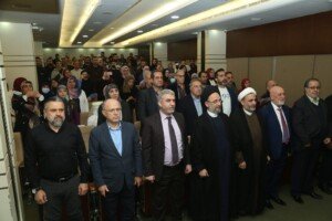 مسؤولون ورجال دين وحشد من الحاضرين يقفون في قاعة مستشفى بهمن خلال زيارة تضامنية مع الجرحى | Officials, clergy, and a crowd of attendees standing in Bahman Hospital's hall during a solidarity visit to the wounded
