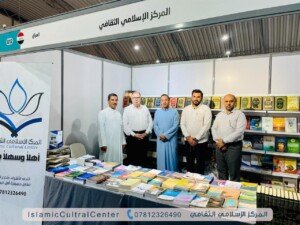 خمسة رجال يقفون أمام جناح يحمل لافتة المركز الإسلامي الثقافي وعلى الطاولة كتب متنوعة | Five men standing in front of a stand bearing the Islamic Cultural Center sign, with various books on the table