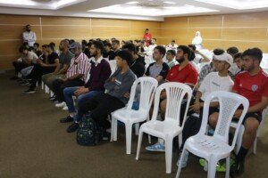 عشرات الشبان يجلسون على كراسي بيضاء في قاعة داخلية يتابعون توجيهات | Dozens of young men sitting on white chairs in an indoor hall following instructions