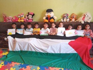 عشرة أطفال يجلسون أمام أريكة يحملون رسومات ملونة فوق علم فلسطين ممدود، وخلفهم دمى محشوة | Ten children sit in front of a couch holding colorful drawings above a spread-out Palestinian flag, with stuffed toys behind them