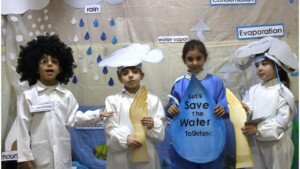 أربعة تلامذة يرتدون أزياء ترمز لعناصر دورة الماء مثل المطر والتبخر ويحملون لافتة Let's Save The Water Together | Four students wearing costumes representing water-cycle elements such as rain and evaporation, holding a sign reading 'Let's Save The Water Together'