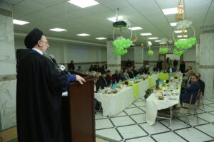 رجل دين يتحدث خلف منصة في قاعة مزينة ببالونات خضراء وبيضاء أمام موائد طويلة للحضور | A cleric speaking behind a podium in a hall decorated with green and white balloons, in front of long tables of guests