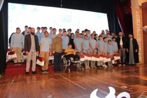 مجموعة كبيرة من الطلاب بالزي المدرسي يقفون على المسرح حاملين أكياس هدايا، وإلى جانبهم رجال دين ومسؤولون، في مشهد يشبه حفل تكليف الطلاب لا لقاء لخريجي الشهادات المهنية | A large group of students in school uniforms stand on stage holding gift bags, alongside clerics and officials, in a scene resembling the students' coming-of-age ceremony rather than a gathering of vocational-certificate graduates