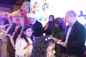 رجل دين ورجل آخر يقفان قرب فتاة صغيرة وسط مجموعة من النساء والأطفال أمام شاشة تعرض صور أطفال في قاعة مزينة للعيد | A cleric and another man stand near a young girl amid a group of women and children in front of a screen showing children's photos, in an Eid-decorated hall