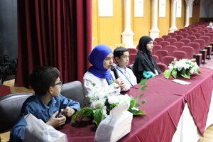 ثلاثة تلامذة يجلسون خلف طاولة عنابية مزينة بالورد على مسرح فارغ بمقاعد حمراء خلفهم | Three students sit behind a maroon, flower-decorated table on a stage with empty red seats behind them