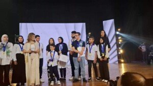 مجموعة طلاب وطالبات يقفون على المسرح بأوسمة تحدي القراءة العربي إلى جانب متحدثتين | A group of students stand on stage wearing Arab Reading Challenge medals alongside two speakers