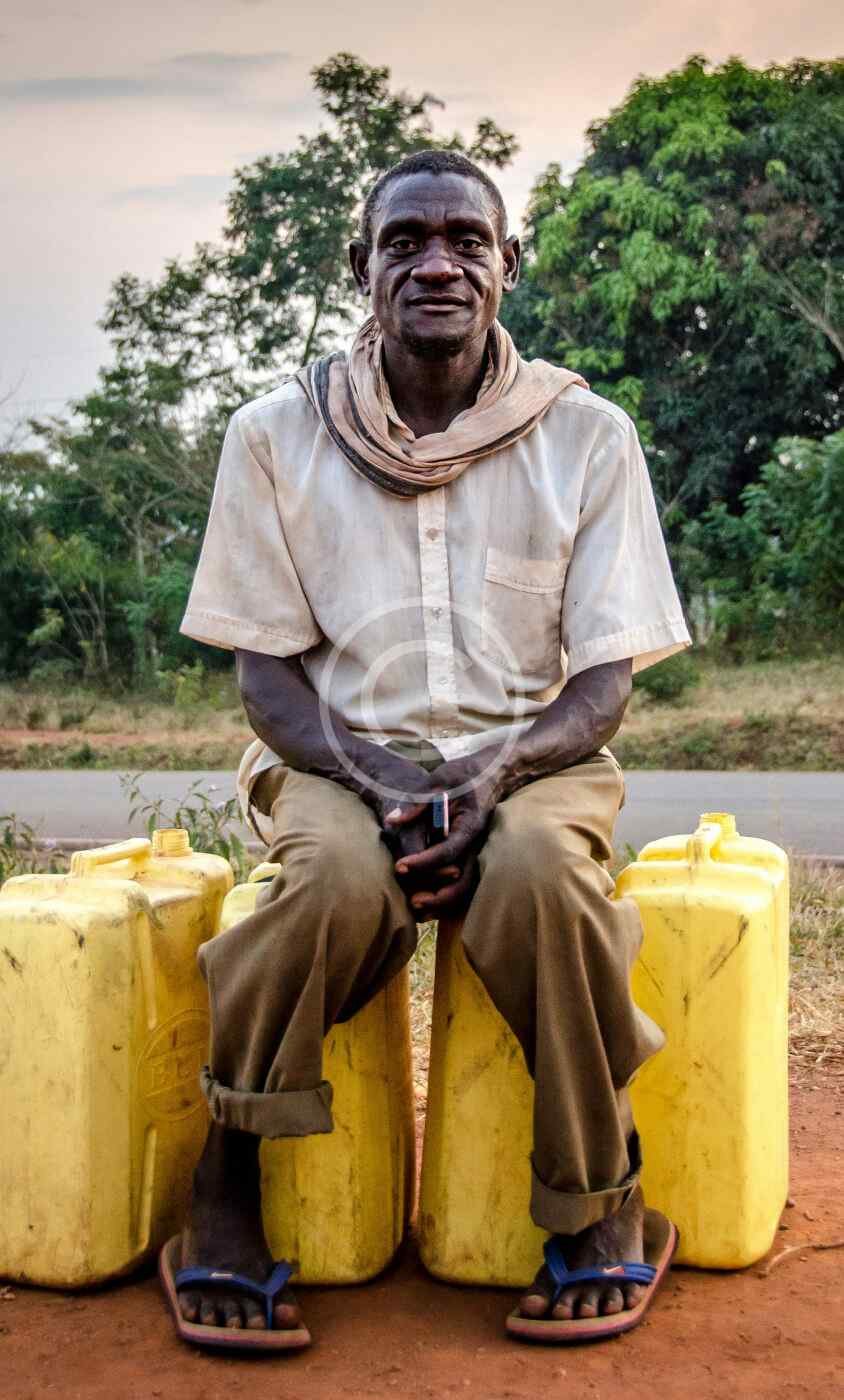 رجل يجلس على عبوات مياه صفراء بجانب طريق ريفي، صورة مخزنة محمية بحقوق نشر | a man sits on yellow water jerrycans beside a rural road, a copyrighted stock photo