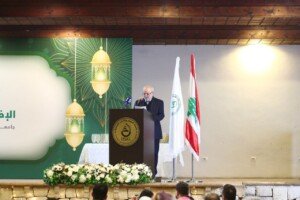 رجل يلقي كلمة خلف منصة USAL أمام علم لبنان ولافتة خضراء لرمضان | A man speaks behind a USAL podium beside the Lebanese flag and a green Ramadan banner
