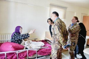 مسنّة بحجاب أرجواني تمد يدها مرحبة بجنديين من اليونيفيل يقفان قرب سريرها في دار الرعاية | An elderly woman in a purple headscarf extends her hand to welcome two UNIFIL soldiers standing near her bed in the care home