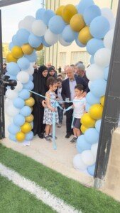 عالم دين وأطفال يقفون تحت قوس بالونات زرقاء وذهبية لحظة قص شريط أزرق | A cleric and children stand under an arch of blue and gold balloons at the moment of cutting a blue ribbon