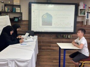 تلميذ يخضع لامتحان شفهي أمام معلمتين ضمن المسابقة الرمضانية لحفظ القرآن الكريم للصف السادس، والشاشة الذكية خلفهما تعرض عنوان المسابقة | A pupil takes an oral exam before two examiners in the sixth-grade Ramadan Quran memorization competition, with the topic shown on the smartboard behind them
