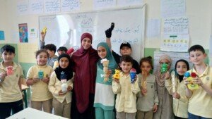 مجموعة تلاميذ وتلميذات يقفون مع معلمة حاملين دمى مصنوعة يدوياً أمام سبورة كتب عليها كلمة الإبداع | A group of pupils stand with a teacher holding handmade puppets in front of a board labeled creativity