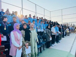 صف من الطلاب يرتدون ميداليات يقفون مع طالبات ومعلمات في ساحة المدرسة عند الغروب | A row of students wearing medals stand with girls and teachers in the school yard at sunset