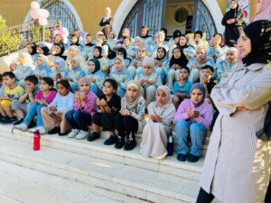 أطفال يجلسون على الدرج رافعين أكفّهم بالدعاء بإشراف معلّمة خلال البرنامج الرمضاني لعوائل الأيتام في البقاع الأوسط | Children sit on the steps raising their palms in prayer, supervised by a teacher, during the Ramadan program for orphan families in central Bekaa