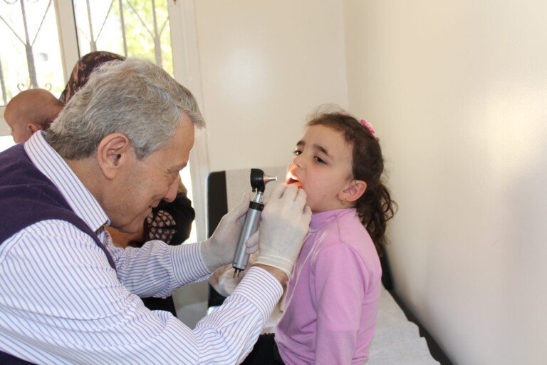 طبيب يفحص فم طفلة بأداة مضيئة في مركز المصطفى الصحي | A doctor examining a little girl's mouth with a lighted instrument at Al-Mustafa Health Center