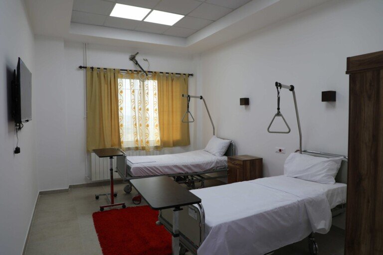 غرفة إقامة بسريرين طبيين مع تلفزيون وستائر صفراء وسجادة حمراء في مركز الرأفة الصحي | A two-bed inpatient room with a wall TV, yellow curtains and a red rug at Al-Ra'fa Health Center
