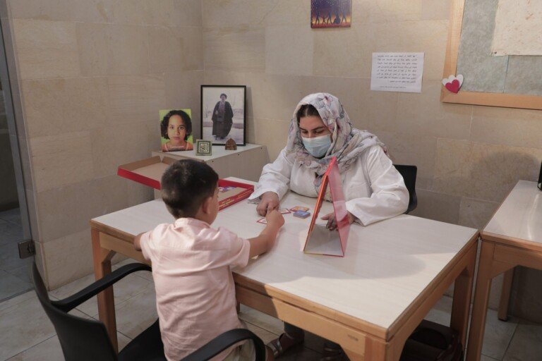 اختصاصية بكمامة تجلس مع طفل أمام بطاقات ولعبة تعليمية في مركز سراج | A masked specialist with a child before cards and an educational game at Siraj Center