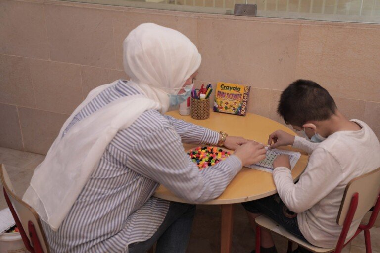 اختصاصية تساعد طفلاً على تركيب خرز ملون في لوحة تعليمية بمركز سراج | A specialist helping a child string colored beads on an educational board at Siraj Center