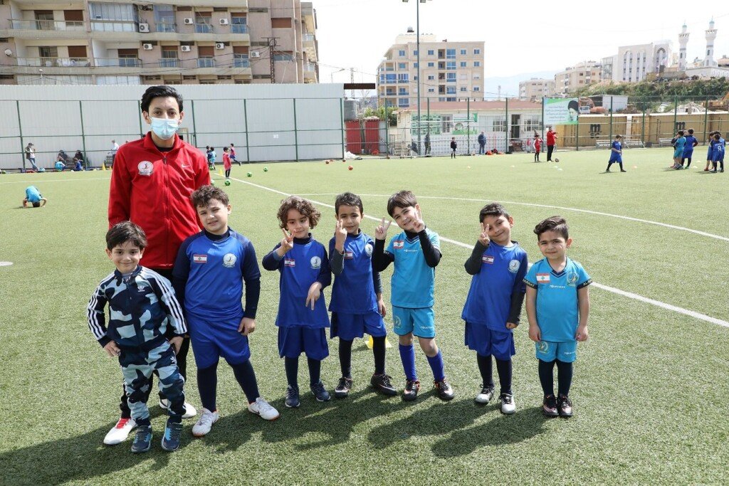 مدرب مع مجموعة من الأطفال بالأزياء الرياضية الزرقاء يقفون على ملعب عشبي في أكاديمية المبرّات الرياضية | A coach with a group of children in blue kits standing on a turf pitch at Al-Mabarrat Sports Academy