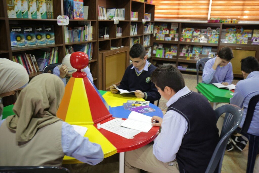 طلاب يشاركون في نشاط قراءة داخل مكتبة مدرسية مع رفوف كتب وطاولة تعليمية | Students participating in a reading activity in a school library with bookshelves and study table