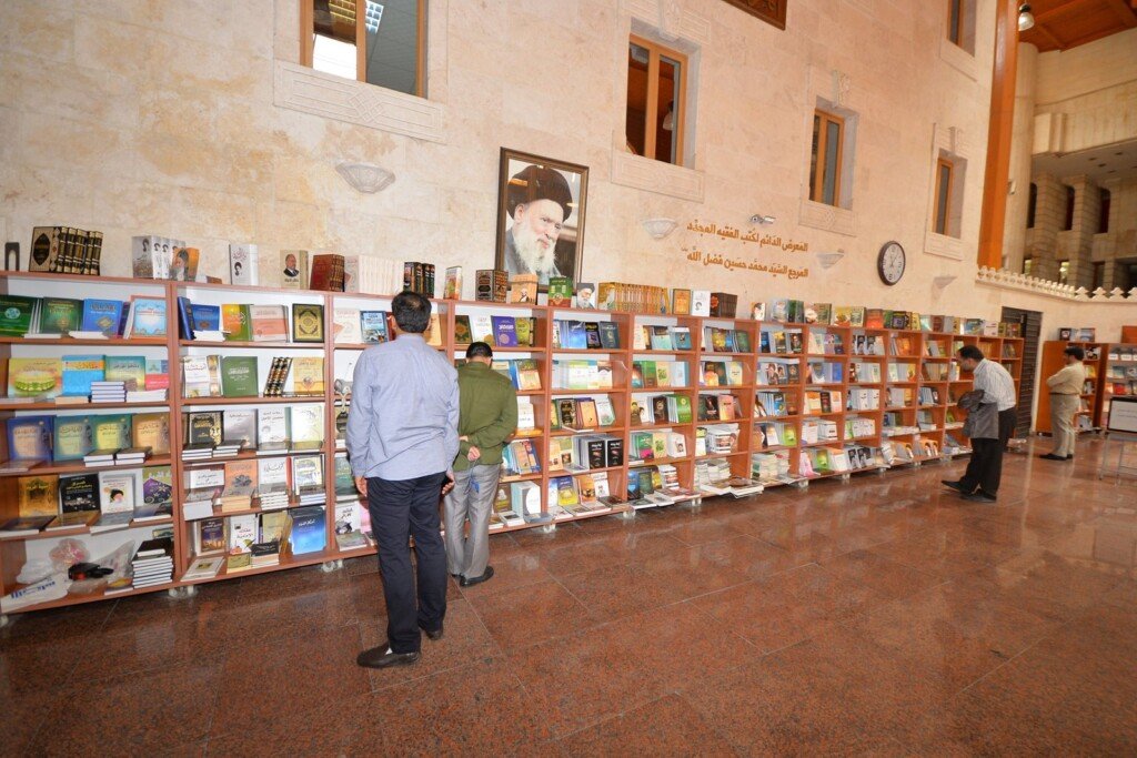 زوار يتصفحون الكتب على رفوف خشبية في معرض دائم للكتب تحت صورة العلامة فضل الله في المركز الإسلامي الثقافي | Visitors browsing books on wooden shelves at a permanent book exhibition beneath a portrait of Allamah Fadlallah at the Islamic Cultural Center