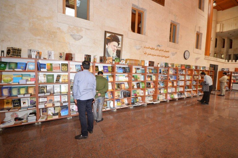 زوار يتصفحون الكتب على رفوف خشبية في معرض دائم للكتب تحت صورة العلامة فضل الله في المركز الإسلامي الثقافي | Visitors browsing books on wooden shelves at a permanent book exhibition beneath a portrait of Allamah Fadlallah at the Islamic Cultural Center