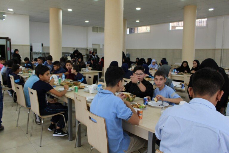 طلاب يجلسون حول طاولات الطعام في مبرّة الحوراء (ع) في لبنان أثناء تناول وجبة ضمن بيئة تعليمية ورعاية اجتماعية | Students sitting around dining tables at Al Hawraa Care Institution in Lebanon during a meal in an educational and social care environment