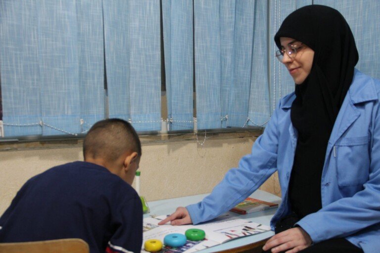 أخصائية بنظارة وحجاب أسود وسترة زرقاء تجلس مقابل صبي وتتابع نشاطاً تعليمياً بأقراص ملوّنة وكتاب على طاولة أمام ستائر زرقاء في مركز HDIC | A specialist with glasses, a black headscarf and a blue jacket sits across from a boy and follows an educational activity with colored discs and a book on a table before blue curtains at the HDIC center