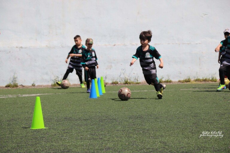 أطفال بزي رياضي مخطط يتدربون على المراوغة والركض حول الشواخص على ملعب عشبي مشمس | Children in striped kits practicing dribbling and running around cones on a sunny grass pitch