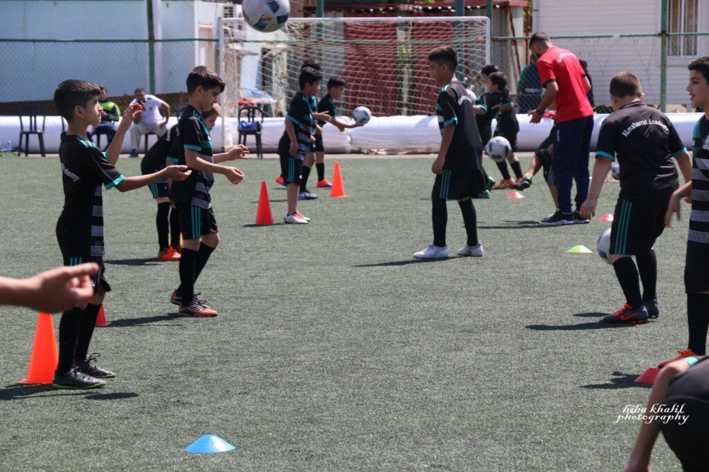 مجموعة من الأطفال والمدربين يؤدون تمارين تمرير الكرات بين الشواخص على ملعب عشبي | A group of children and coaches performing ball-passing drills among cones on a grass pitch