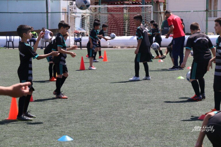 مجموعة من الأطفال والمدربين يؤدون تمارين تمرير الكرات بين الشواخص على ملعب عشبي | A group of children and coaches performing ball-passing drills among cones on a grass pitch