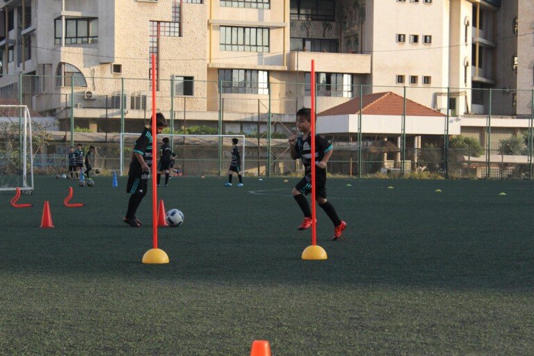طفلان يتدربان على المراوغة حول شواخص برتقالية وكرة قدم على ملعب عشبي أمام مبانٍ سكنية | Two boys practicing dribbling around orange poles with a football on a grass pitch in front of residential buildings