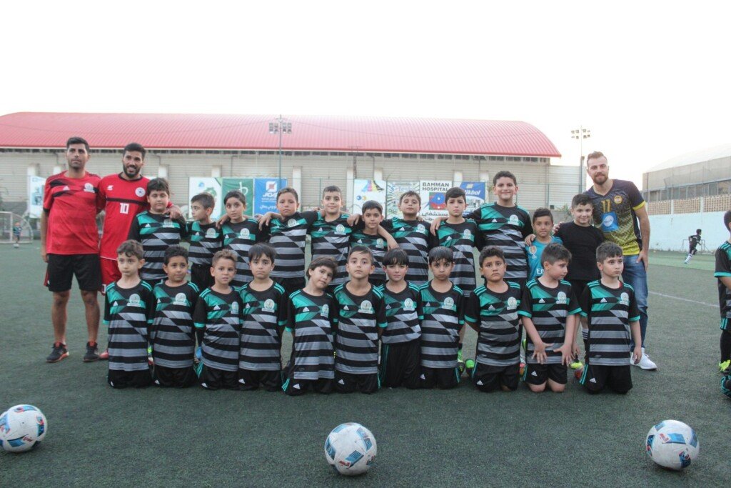 فريق من الأطفال بالزي الرياضي المخطط مع مدربيهم في صورة جماعية على الملعب مع كرات القدم | A team of children in striped kits with their coaches in a group photo on the pitch with footballs