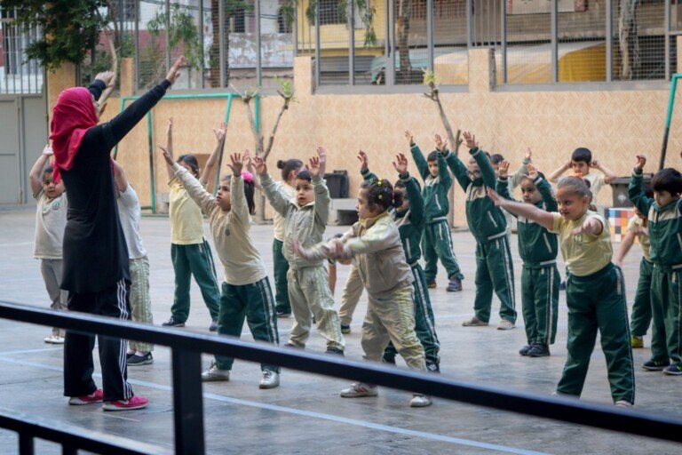 طلاب يشاركون في تمارين رياضية جماعية بإشراف المعلمة داخل ساحة المدرسة | Students participating in group physical exercises under teacher supervision in a school courtyard