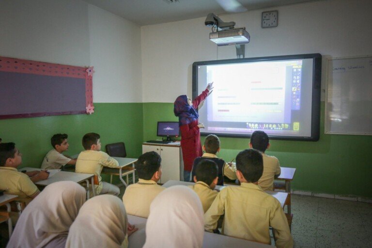 معلمة تشرح درساً باستخدام شاشة ذكية تفاعلية أمام الطلاب داخل صف مدرسي | Teacher explaining a lesson using an interactive smart board in front of students inside a classroom