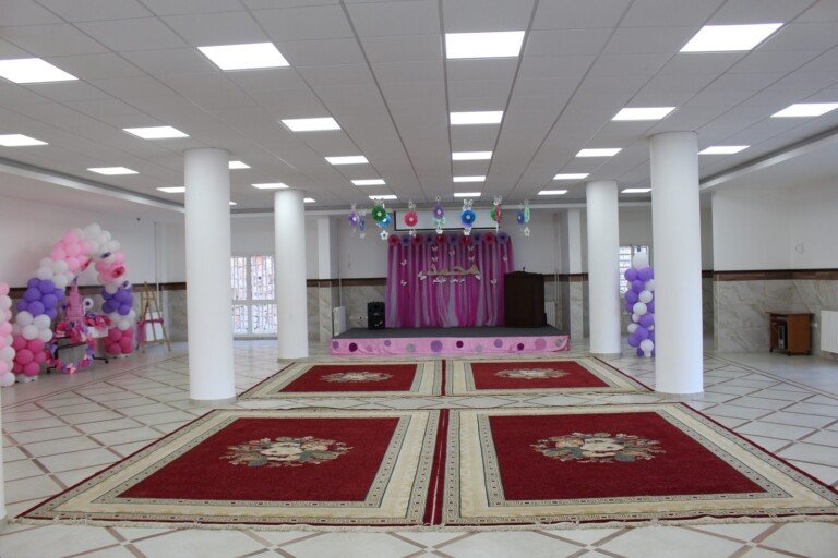 قاعة واسعة مزينة بمنصة احتفالات وسجاد مزخرف لاستقبال الفعاليات في مبرة الإمام الرضا (ع) في لبنان | Spacious decorated hall with a stage and carpets for events at Imam Al-Ridha Institution in Lebanon