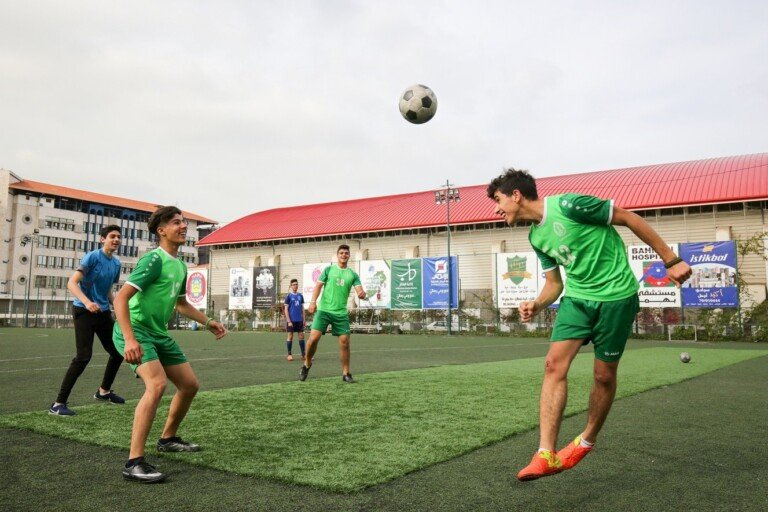 فتيان يلعبون كرة القدم على ملعب عشبي أحمر السقف ضمن أنشطة دار الصادق للتربية والتعليم | Boys playing football on a turf pitch with a red-roofed hall, part of Dar Al-Sadiq for Education activities