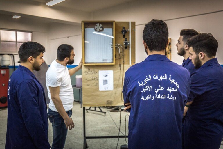 طلاب يرتدون بزّات زرقاء مكتوب عليها معهد علي الأكبر ورشة التدفئة والتبريد يتابعون شرحاً حول نموذج تبريد معلّق أمامهم في المعهد | Students in blue overalls printed with Ali Al-Akbar Institute heating and cooling workshop follow an explanation of a mounted refrigeration model in front of them at the institute